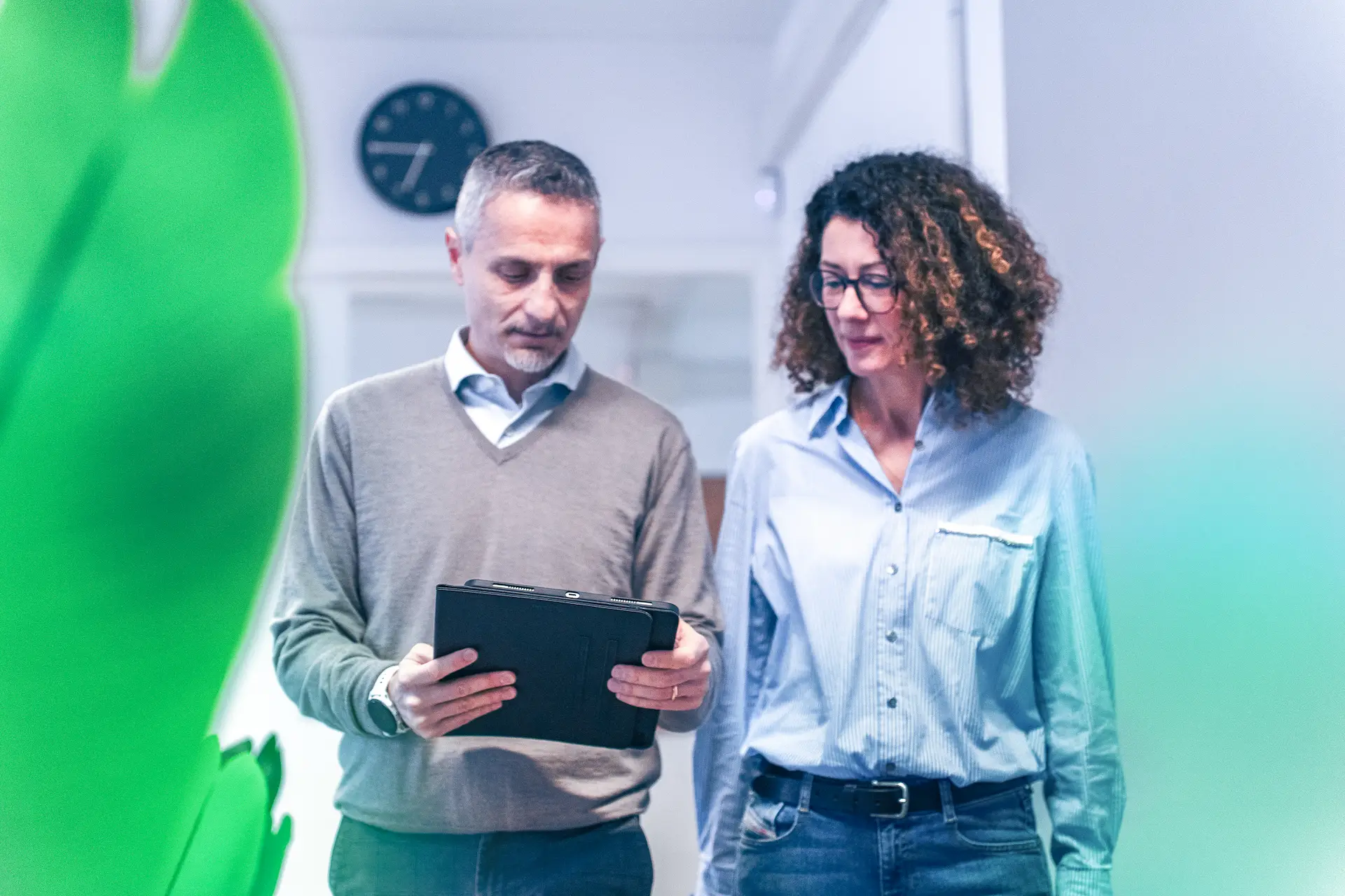 Due professionisti di Winet discutono di un progetto di ricerca e sviluppo, consultando un tablet in un corridoio luminoso con un orologio sullo sfondo.