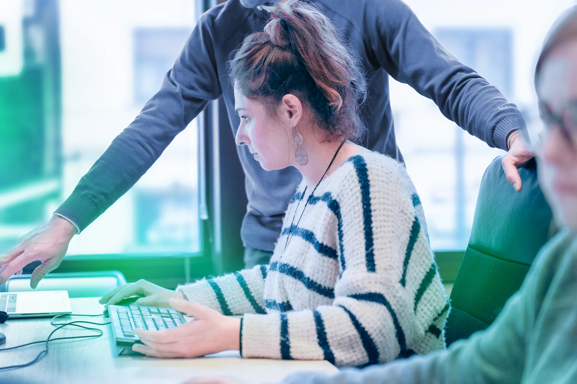 Una donna con un maglione a righe è concentrata al computer, mentre un collega in piedi alle sue spalle indica qualcosa sullo schermo in un ufficio luminoso.