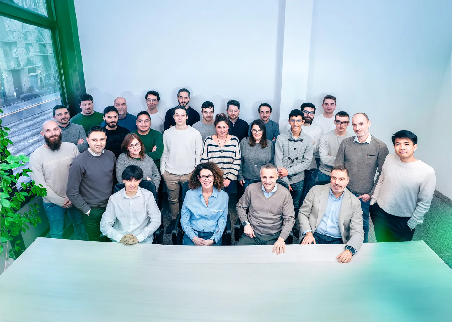 Foto di gruppo di un team aziendale composto da uomini e donne di varie età; alcuni seduti attorno a una scrivania, altri in piedi dietro di loro, tutti sorridenti in un ufficio luminoso con piante e grandi finestre sullo sfondo.