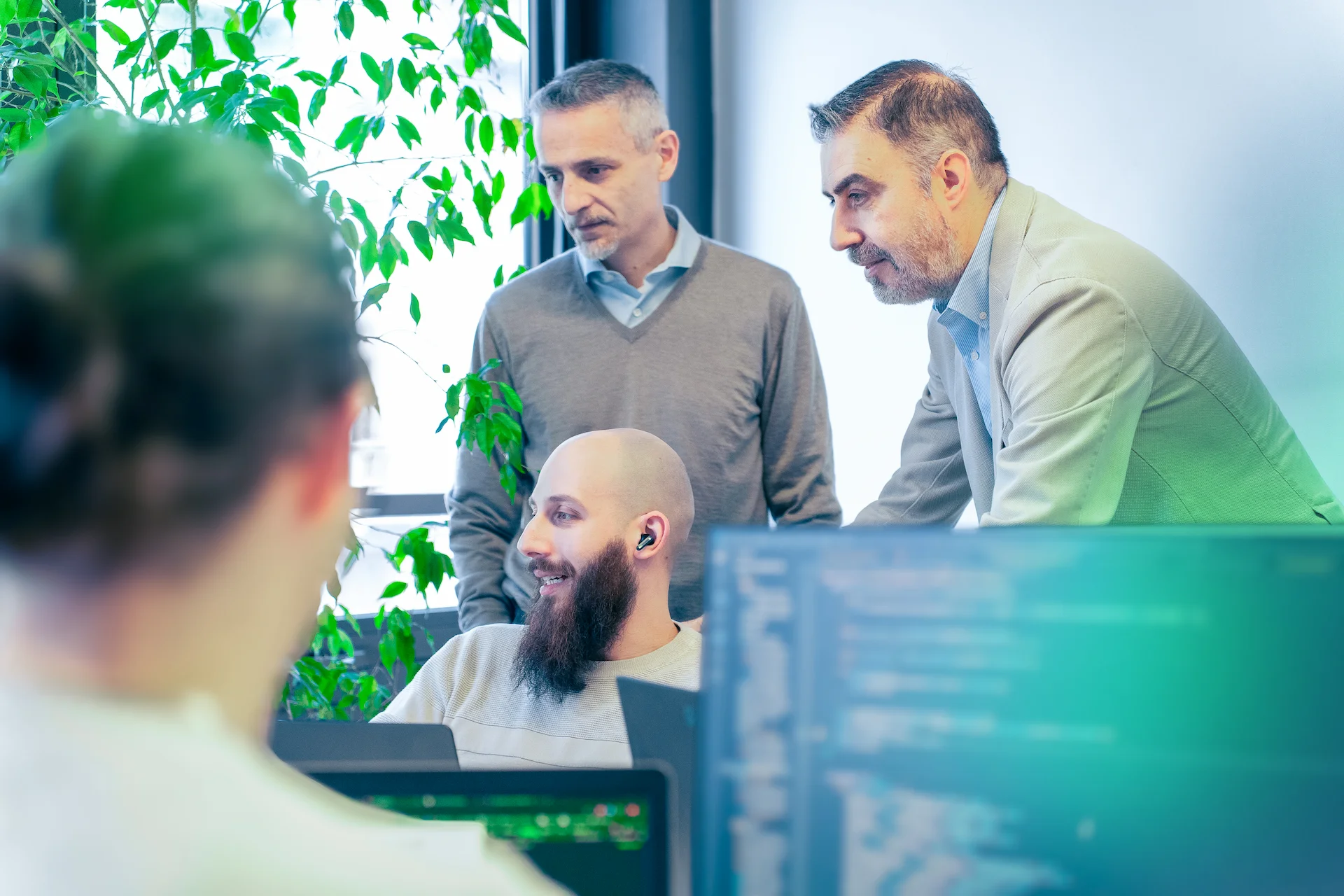 Un uomo seduto con auricolari e barba sorride mentre due colleghi in piedi gli parlano in un ufficio luminoso; sullo sfondo piante verdi e schermi con codice in primo piano.