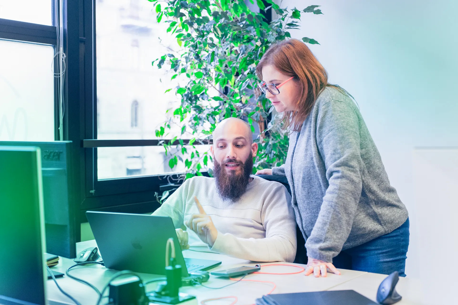 Due collaboratori di Winet discutono di un progetto software per Thales, con un laptop su una scrivania in un ufficio luminoso e una grande pianta sullo sfondo.