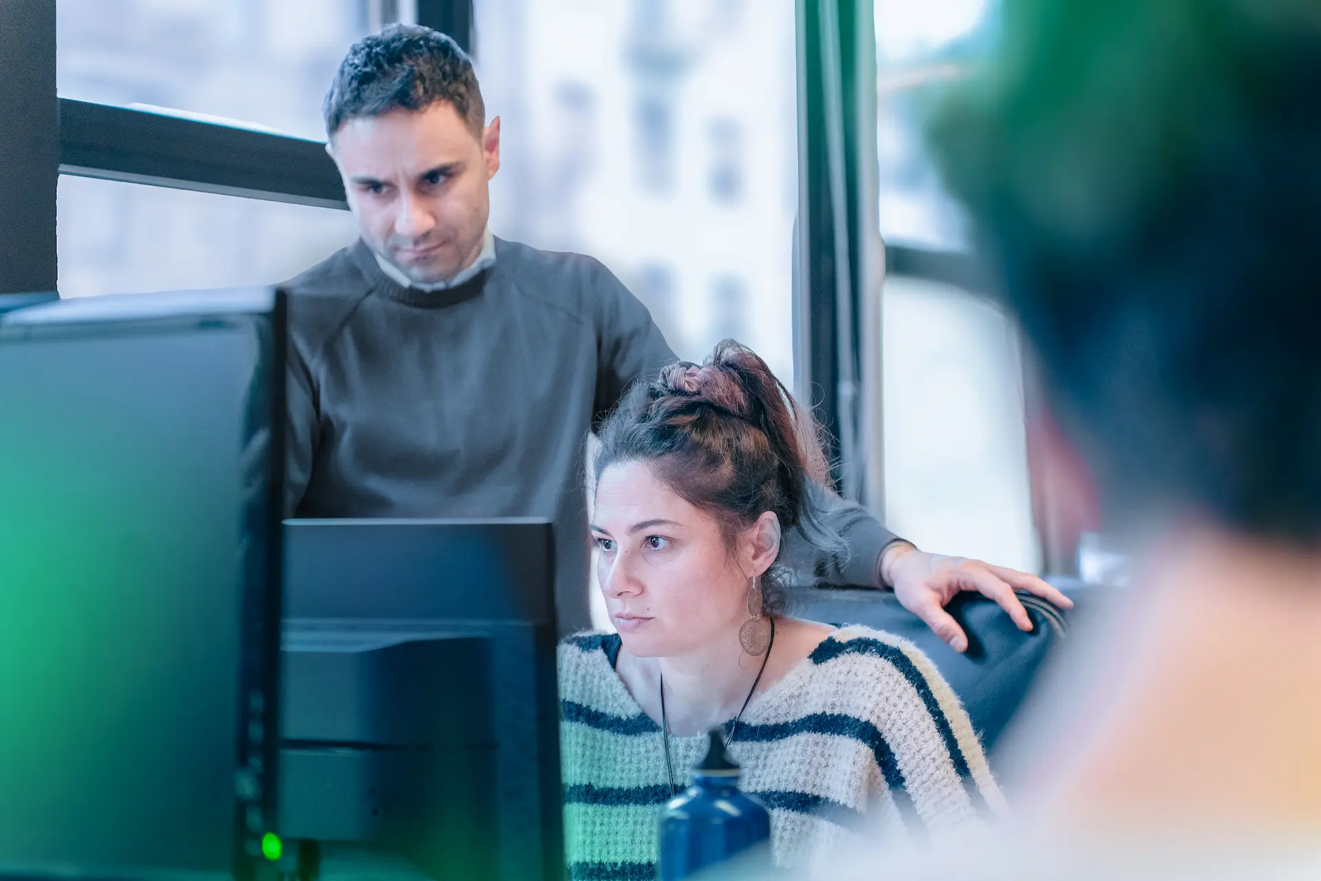 Due collaboratori di Winet al lavoro su un progetto software per Thales: la collega è seduta al computer, concentrata sul monitor, mentre il collega in piedi la assiste in un ufficio luminoso.