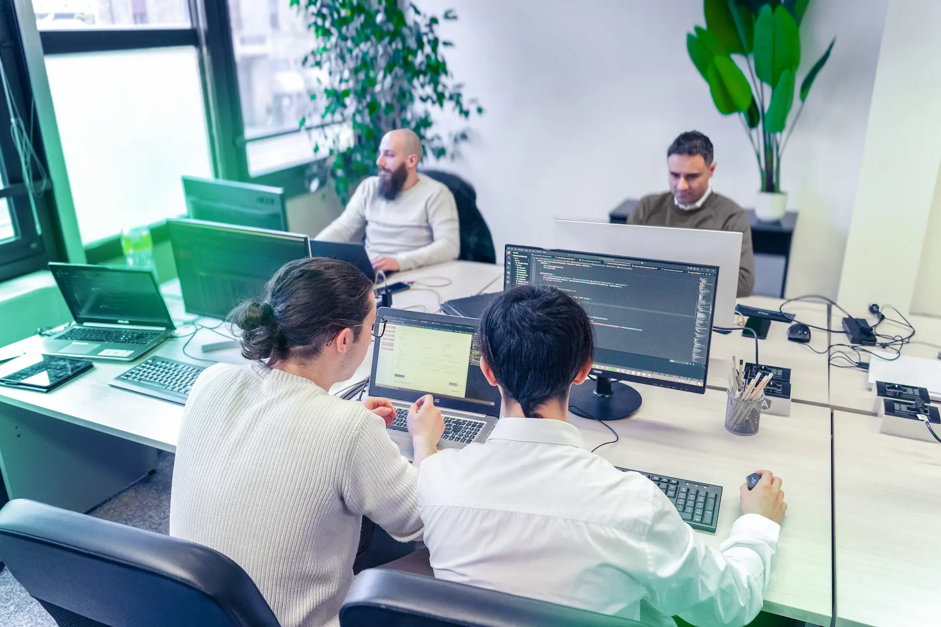 Team di Winet al lavoro su progetti software in un ufficio open space, con due sviluppatori che collaborano davanti a un monitor e altri colleghi impegnati sullo sfondo.