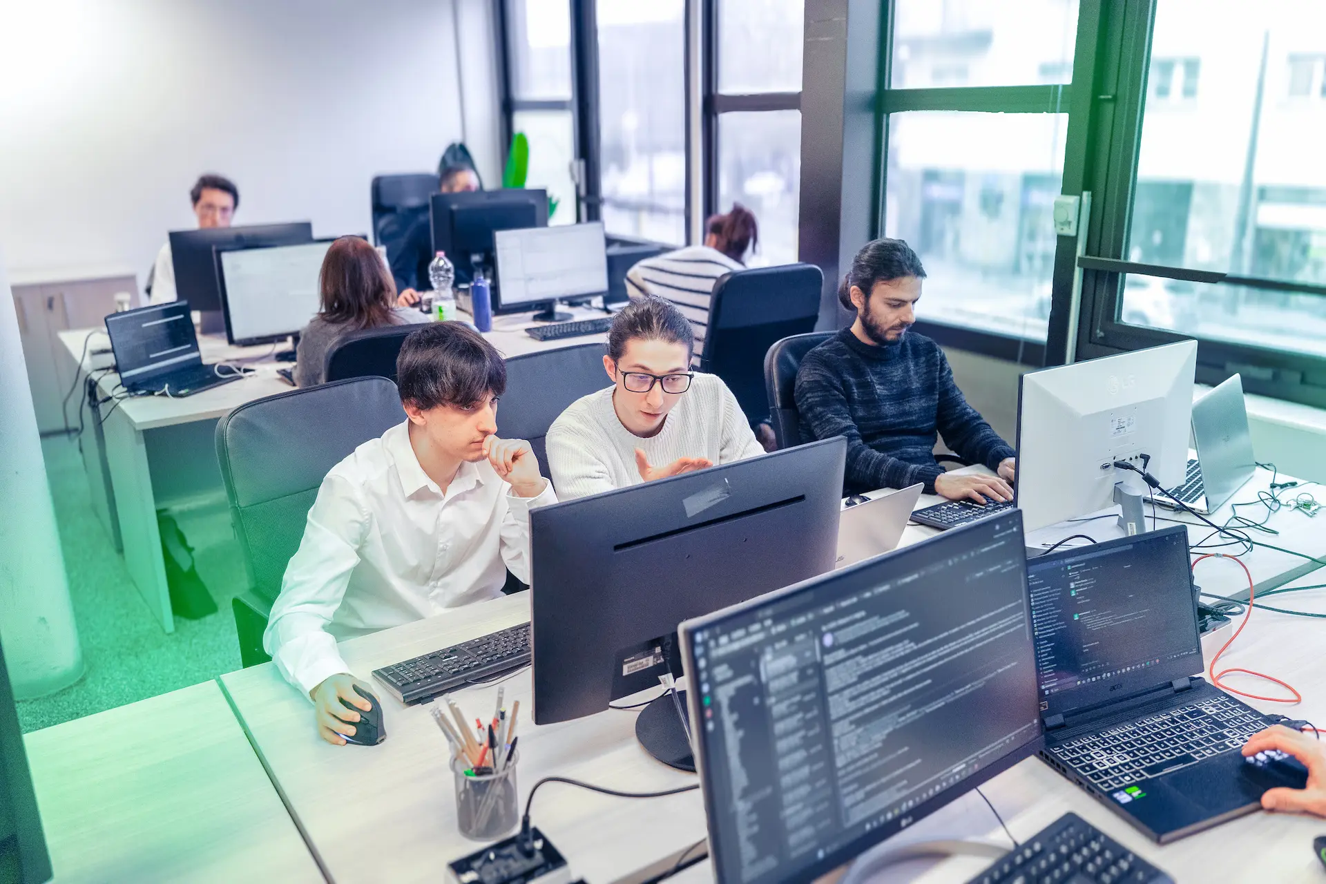 Team di Winet al lavoro in un ufficio open space, con più postazioni informatiche e collaboratori che discutono e scrivono codice su diversi monitor.