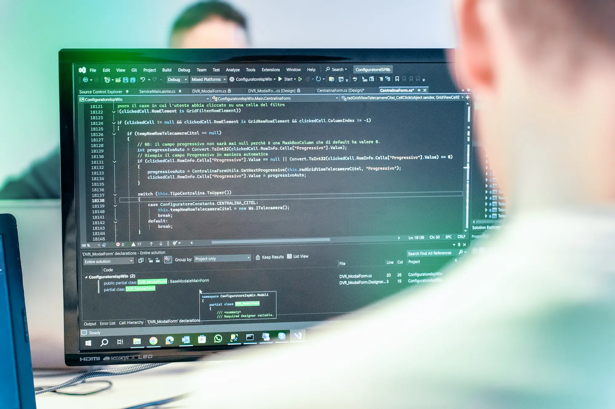 Uno sviluppatore di Winet, ripreso di spalle, controlla il codice sorgente su due monitor, concentrandosi sullo sviluppo e il debugging di un progetto software.