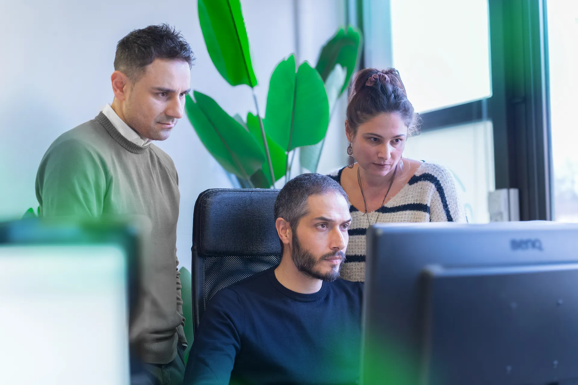 Tre colleghi di un team di sviluppo software, uno seduto al computer e gli altri due in piedi dietro di lui, osservano il monitor con attenzione in un ufficio luminoso.